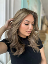 Side angle of model wearing Ashli, a lace top wig in honey brown with soft beige blonde highlights, styled in flowing, beachy waves.