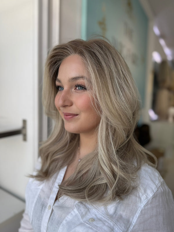Model wearing a long, cool-toned ash blonde lace front wig with realistic hairline and layered volume, styled in soft curls.