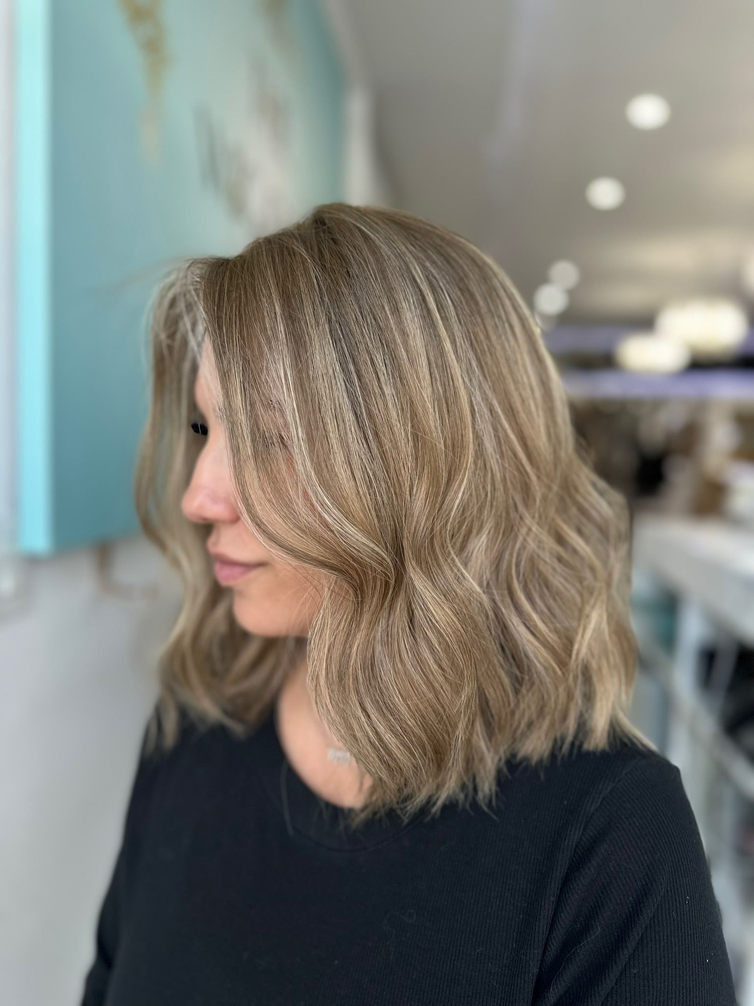 Wavy side angle view of the Margot wig in sandy beige blonde with ashy lowlights, styled in a soft, 15-inch length.