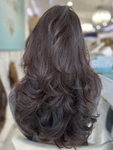 Alana wig by The Wig Fairy, black wavy hair with subtle highlights, worn by a smiling woman in a stylish, modern setting.