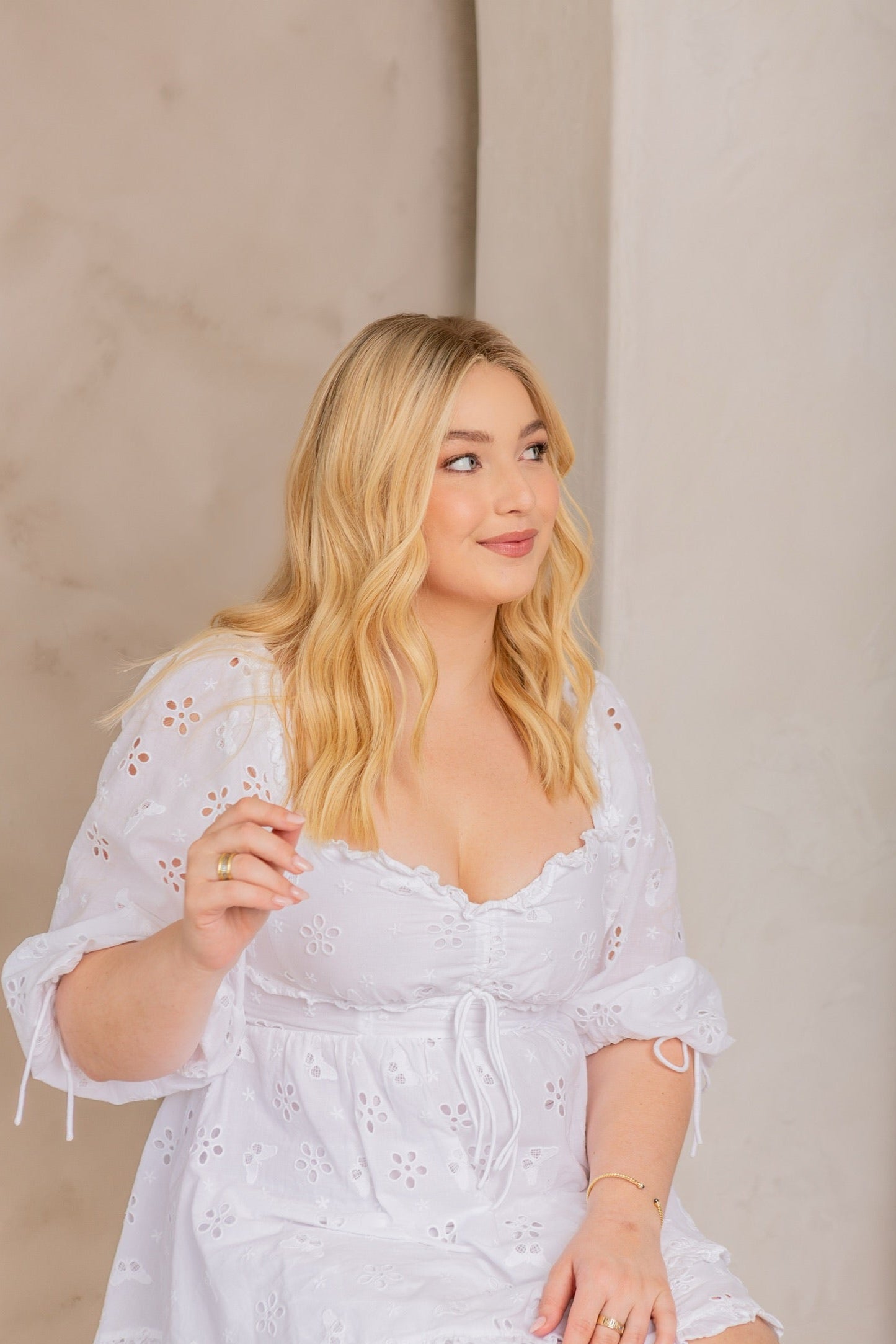 Woman with shoulder-length blonde human hair wig wearing a white embroidered dress, seated against a beige background – Wig Fairy